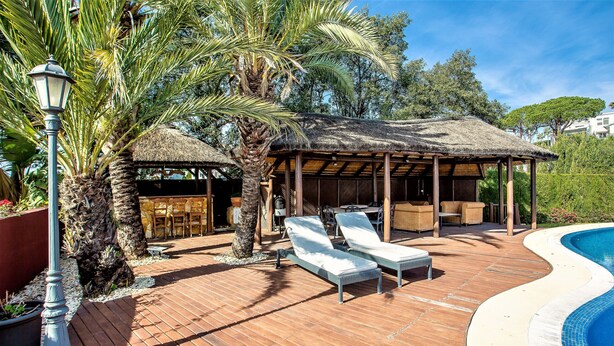 Una terrazza in legno con poltrone relax, un gazebo con tetto di paglia e una piscina.