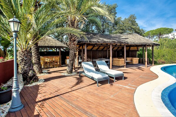 A wooden deck with lounge chairs, a thatched-roof gazebo, and a swimming pool. - Villa Sunsets - charming villa in residential area in Elviria (Marbella)