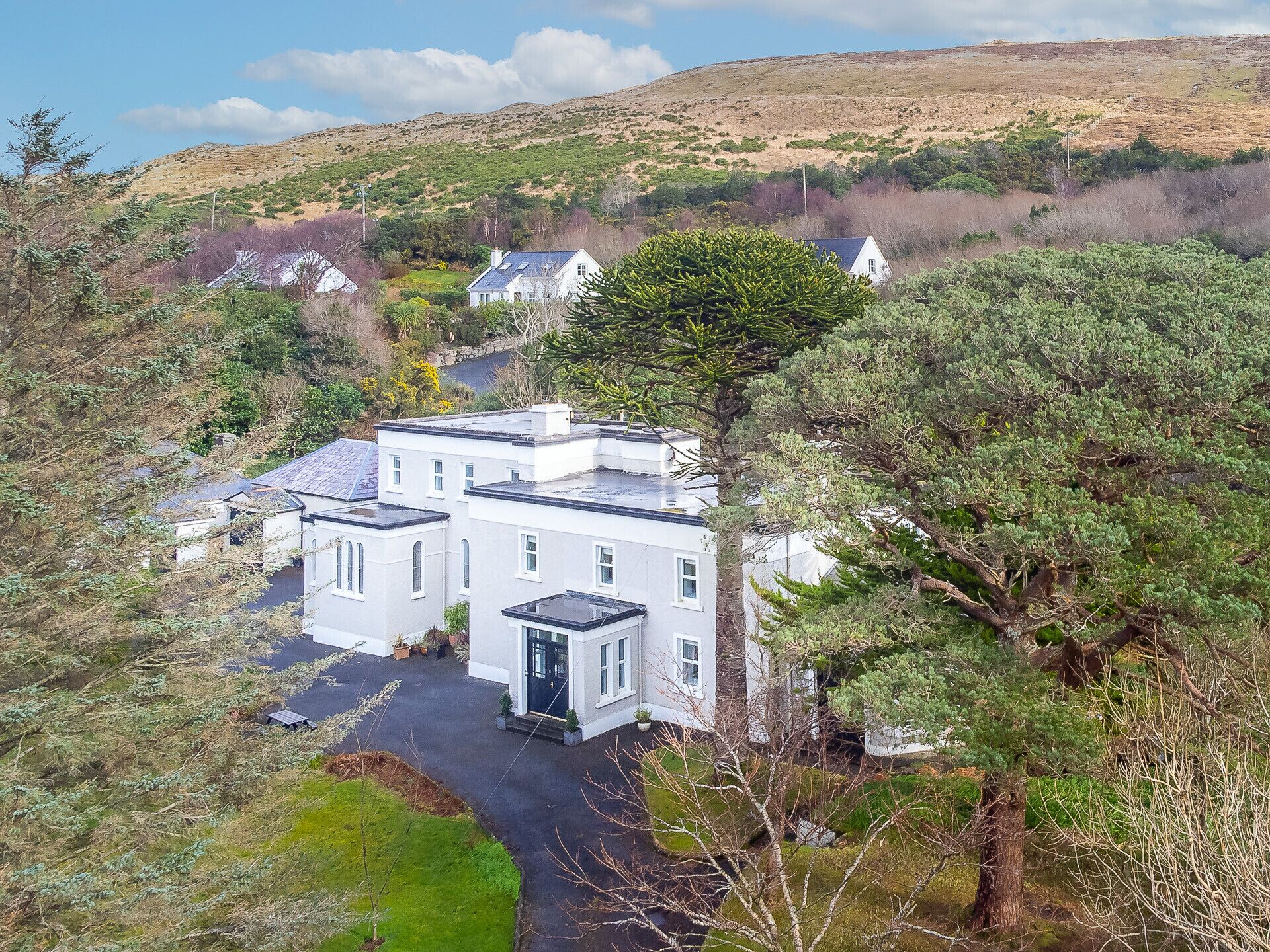 A white building with a flat roof, surrounded by trees and a hill in the background.