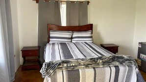 Una camera da letto con un letto, due comodini, una testiera in legno, una finestra con tende e un condizionatore d'aria.