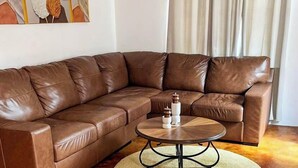 Un divano angolare in pelle marrone, un tavolino rotondo con base metallica e un tappeto dal pelo lungo su un pavimento in legno.