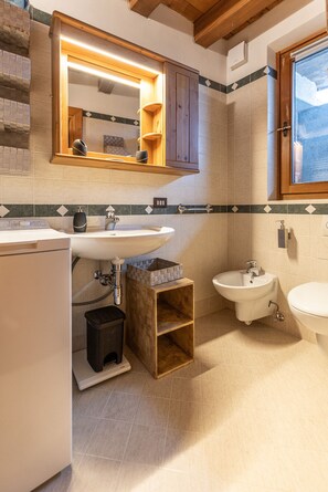 A bathroom with a sink, bidet, and toilet. There is a mirror above the sink and wooden shelves. - Casa Noce, quiet and surrounded by nature (Cortina d´Ampezzo)