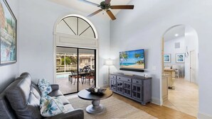 Een woonkamer met een grijze bank, een ronde salontafel en een televisie op een kast. Er is een plafondventilator, een groot raam en een deur naar een patio met tuinmeubilair.