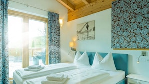 A double bed with white linens and towels, a blue headboard, a nightstand, a wall-mounted lamp, and a painting of a mountain scene. - eder.VIEW (Maria Alm am Steinernen Meer)