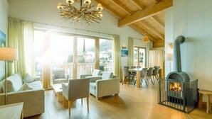 A spacious living room with a high ceiling, a chandelier, a fireplace, and a dining area with a view of the mountains. - eder.VIEW (Maria Alm am Steinernen Meer)