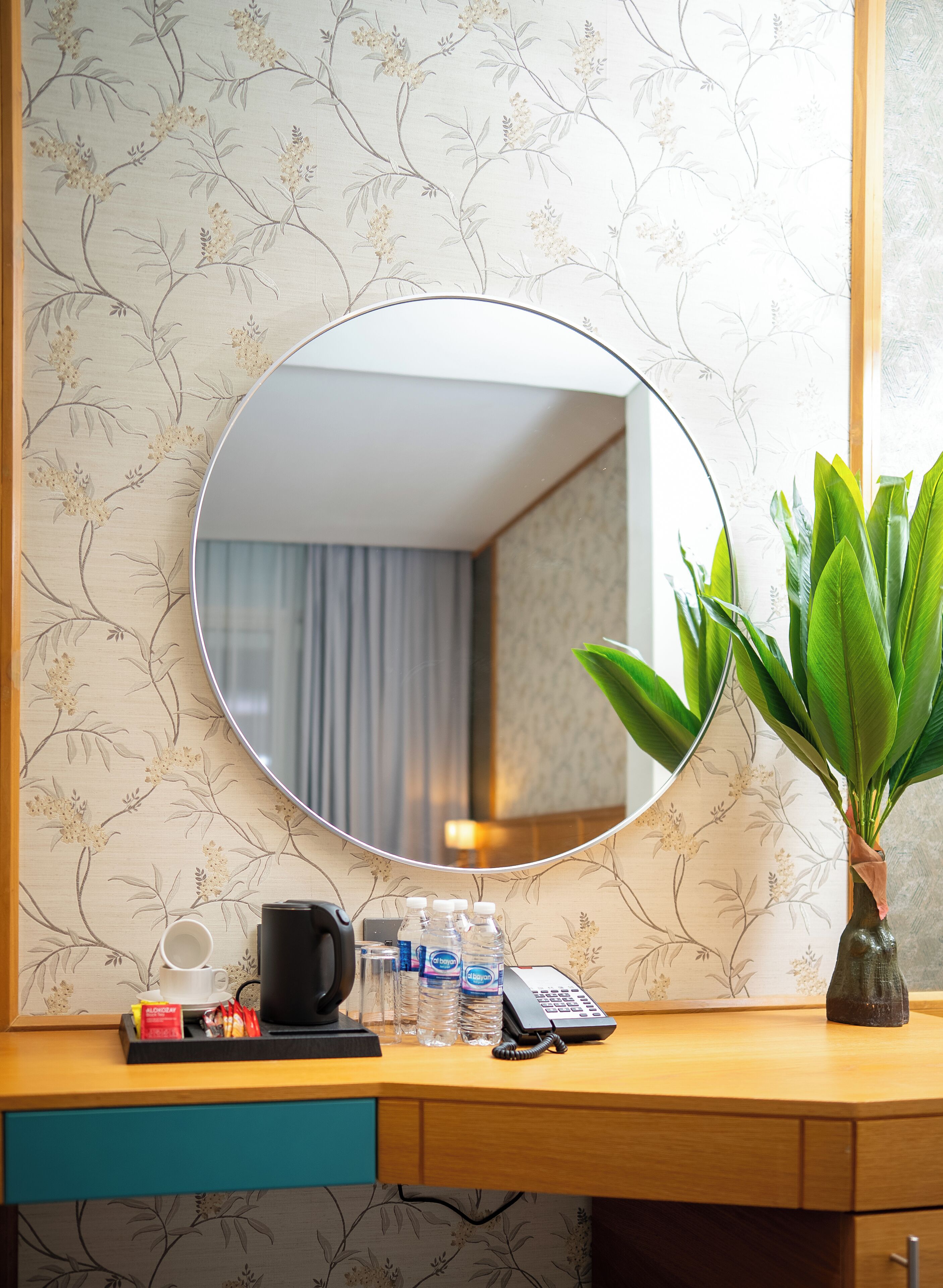Une chambre d’hôtel avec un bureau en bois, un miroir rond, une plante en pot, une machine à café, un téléphone et des bouteilles d’eau.