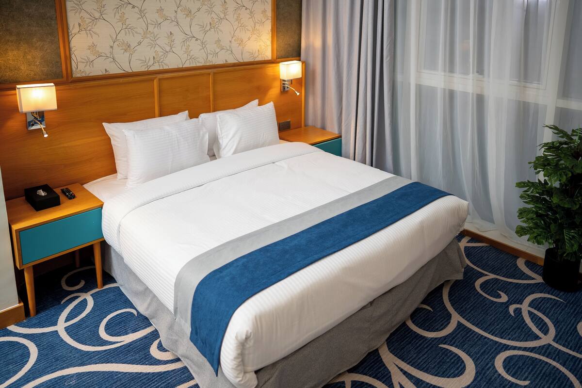 A hotel room with a neatly made bed, two bedside tables with lamps, a potted plant, and a patterned carpet.