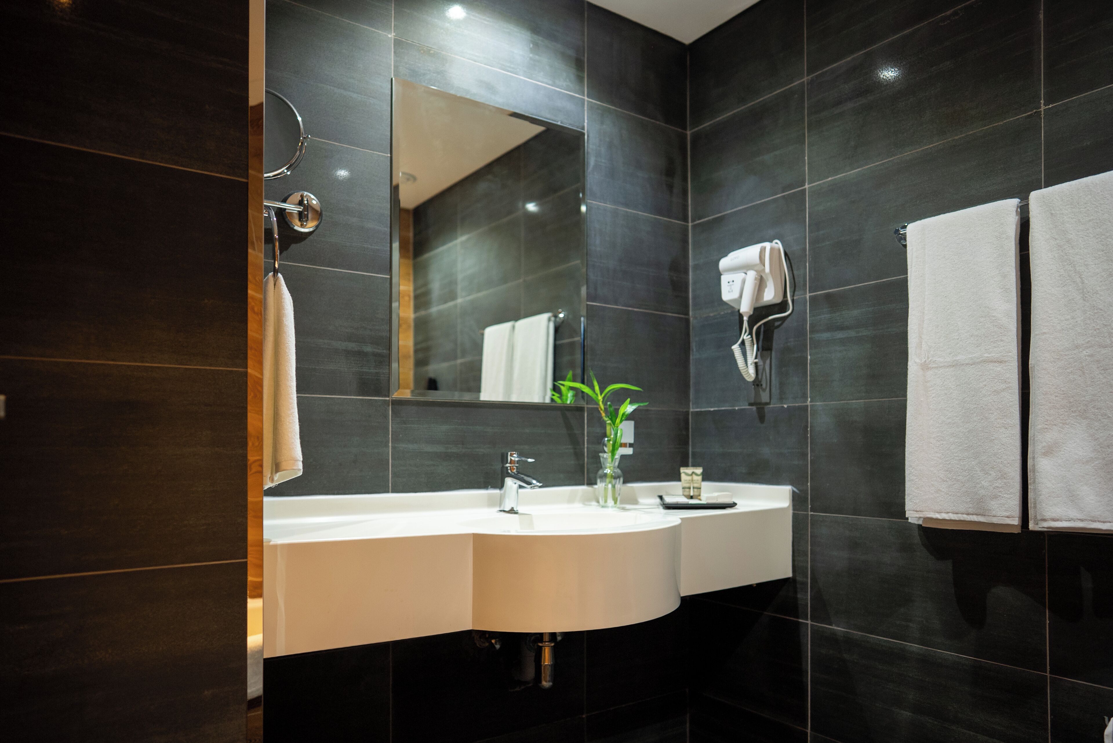 Een moderne badkamer met een donkere betegelde muur, een witte wastafel, een spiegel, een haardroger en twee handdoeken.