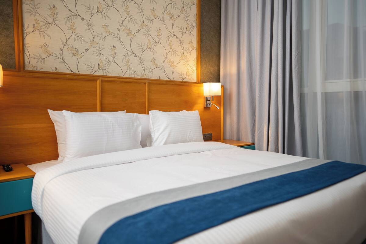 A hotel room with a wooden headboard, a bed with white linens and a blue throw, a nightstand with a lamp, and a wall with floral wallpaper.