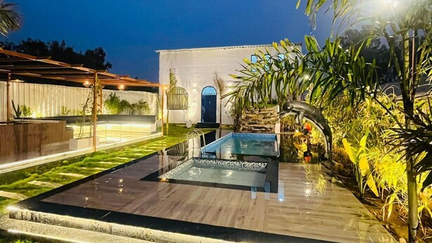 A modern outdoor pool area with a wooden deck, surrounded by lush greenery and a white building in the background. - Greek House By Housiify (Gurugram)