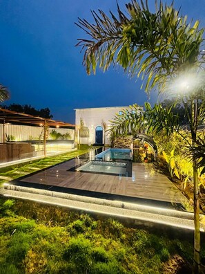 A modern outdoor pool area with a wooden deck, surrounded by lush greenery and a white building in the background. - Greek House By Housiify (Gurugram)