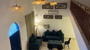 A modern living room with a sectional sofa, a coffee table, and a dining area. There are multiple hanging lights and a large window with a view. - Greek House By Housiify (Gurugram)