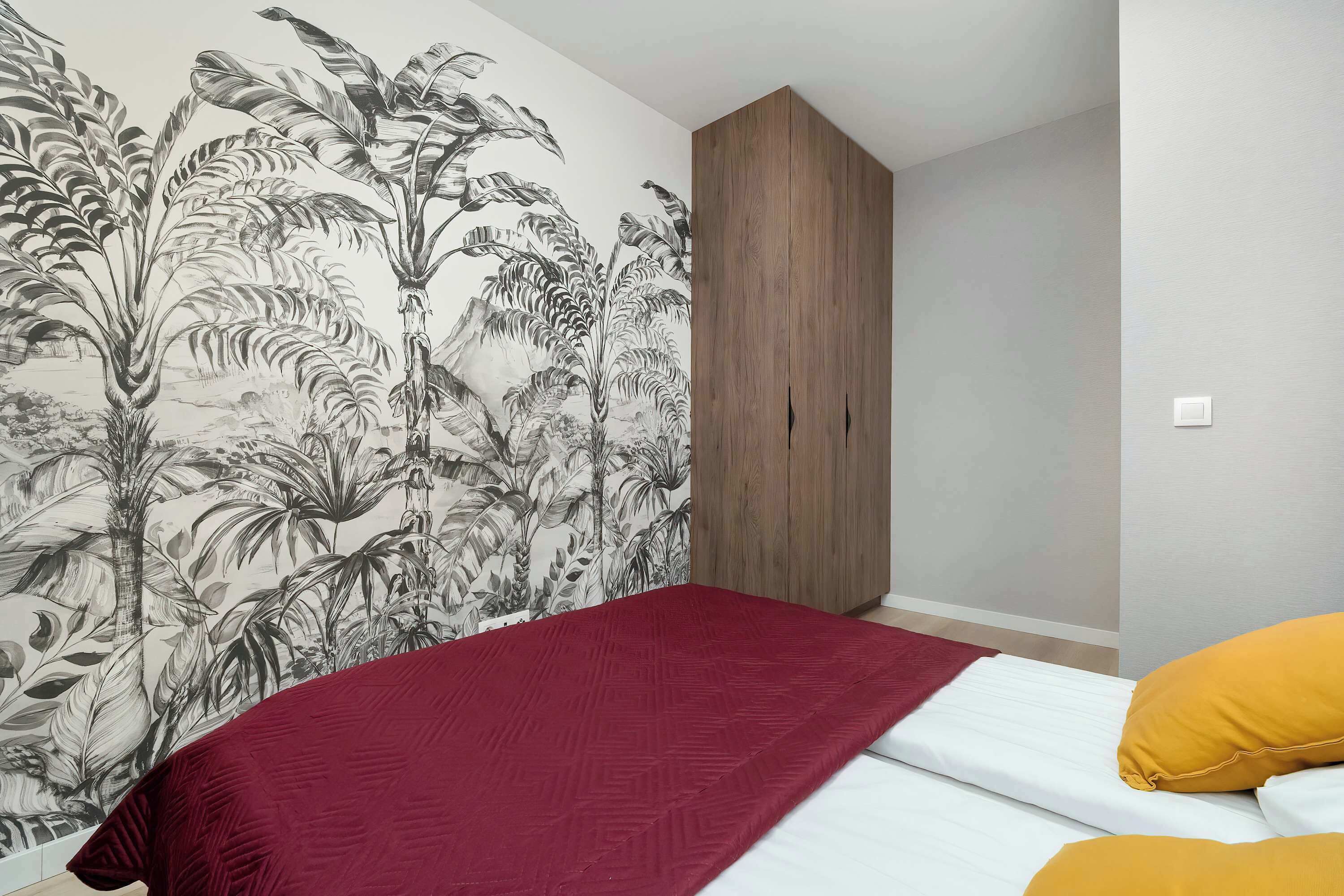 Quarto com cama, manta vermelha e travesseiro amarelo. Há um guarda-roupa e uma parede com um mural de planta tropical.