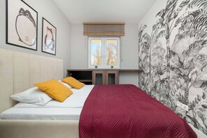 A bedroom with a bed, two pillows, a red blanket, a window with blinds, and a wall with a tropical wallpaper. - Modern Apartment | Balcony, Parking, Close to Expo (Łódź)