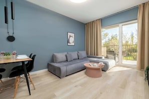 A modern living room with a grey sofa, a round coffee table, and a dining area with a table set for two. - Modern Apartment | Balcony, Parking, Close to Expo (Łódź)