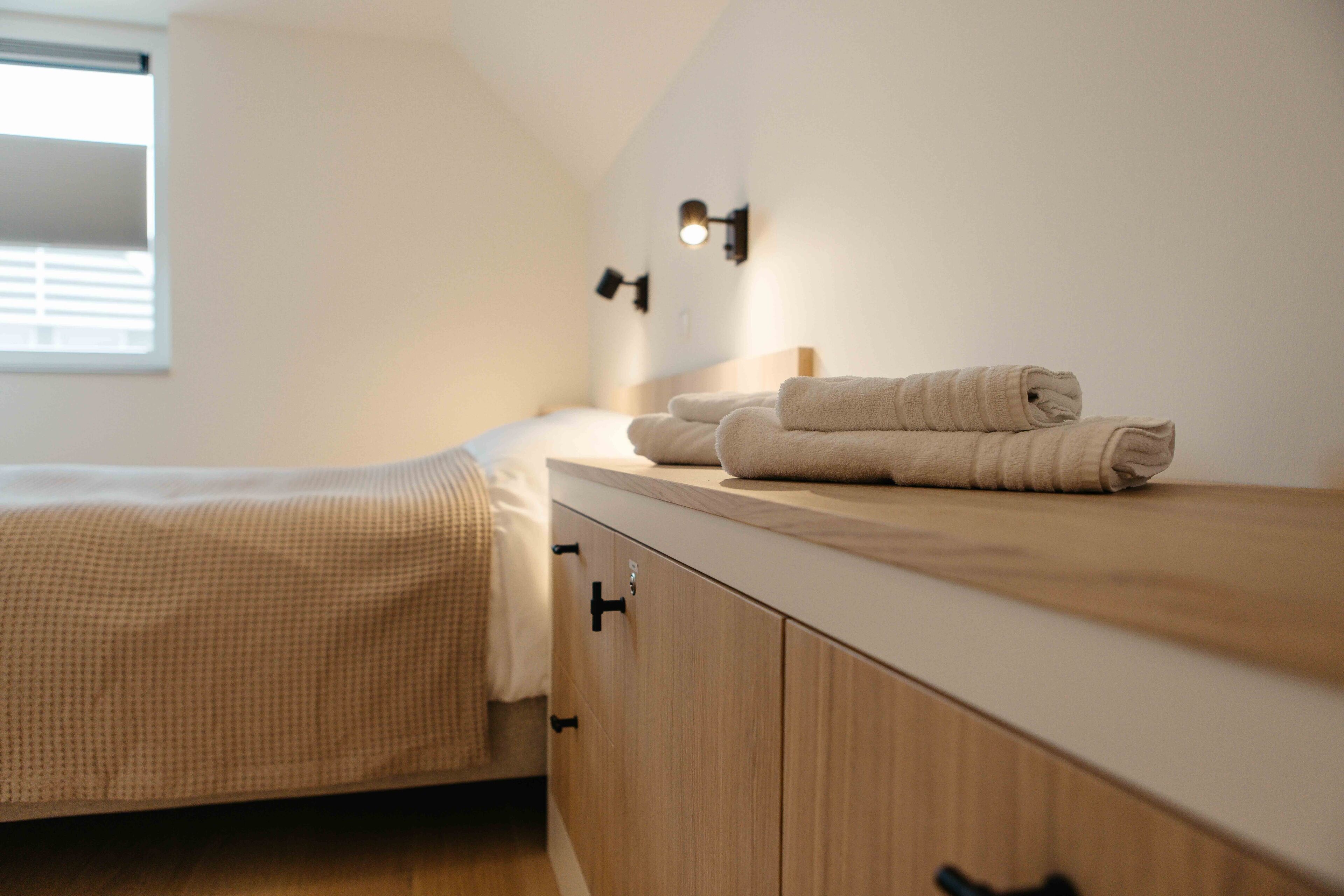 Un lit bien fait, recouvert d’une couverture à carreaux beige, une table de chevet en bois avec des serviettes pliées, et un luminaire fixé au mur.