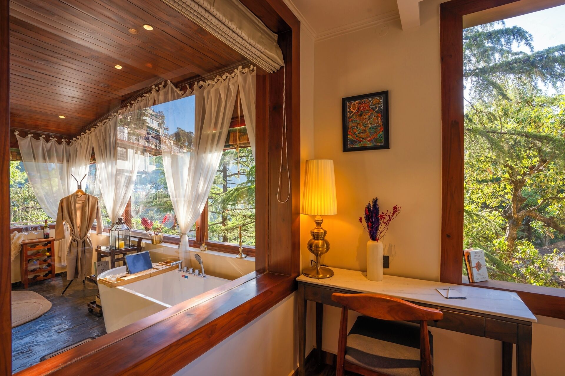 A room with a wooden ceiling, a large window with curtains, a desk with a lamp, a chair, and a view of greenery outside.