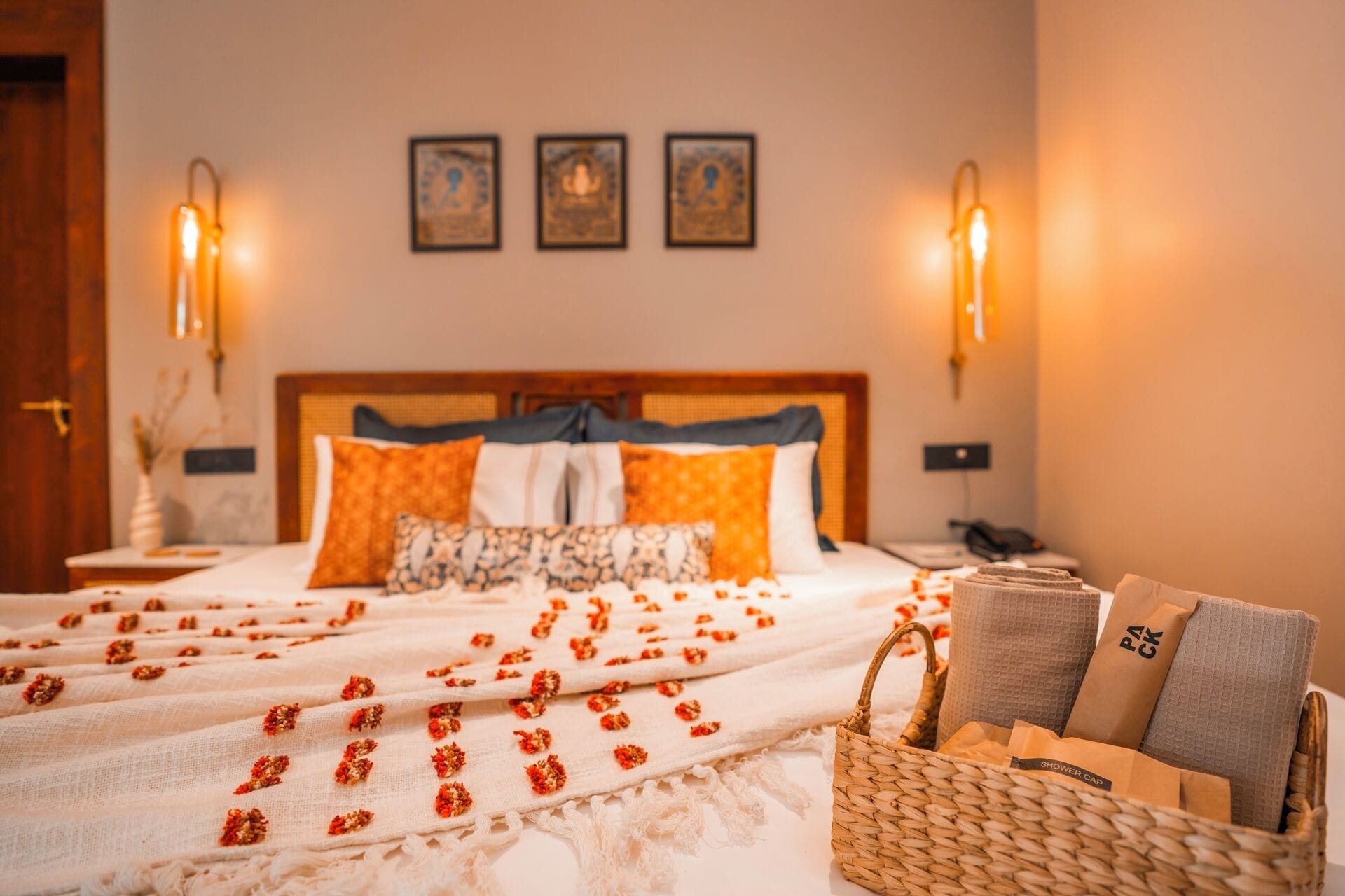 A neatly made bed with a white and orange patterned bedspread, a woven basket with towels and a shower cap, and two wall-mounted light fixtures.