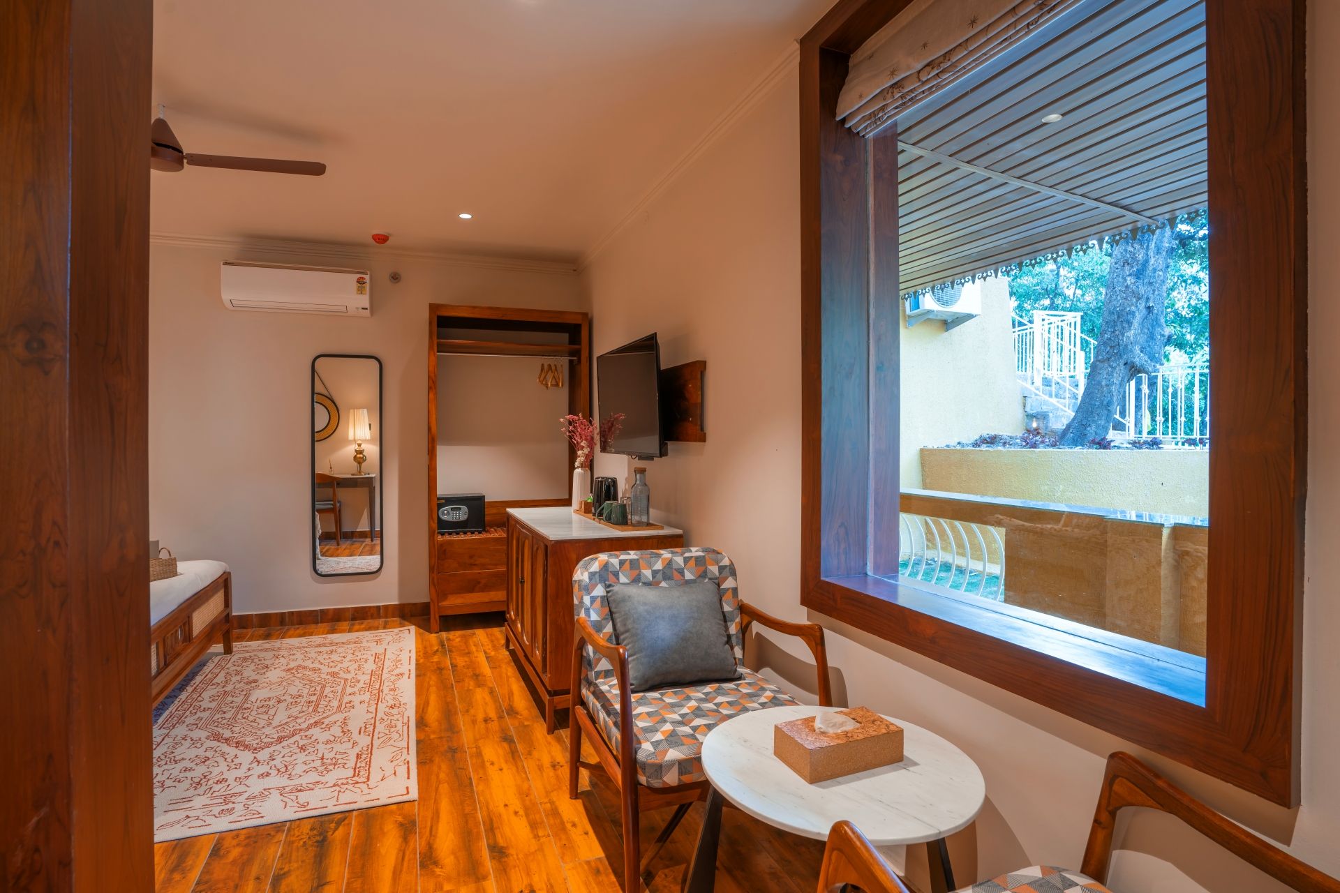 Una stanza con pavimento in legno, un tavolo rotondo, un divano con cuscini decorati, una TV fissata al muro e una finestra vista esterna.