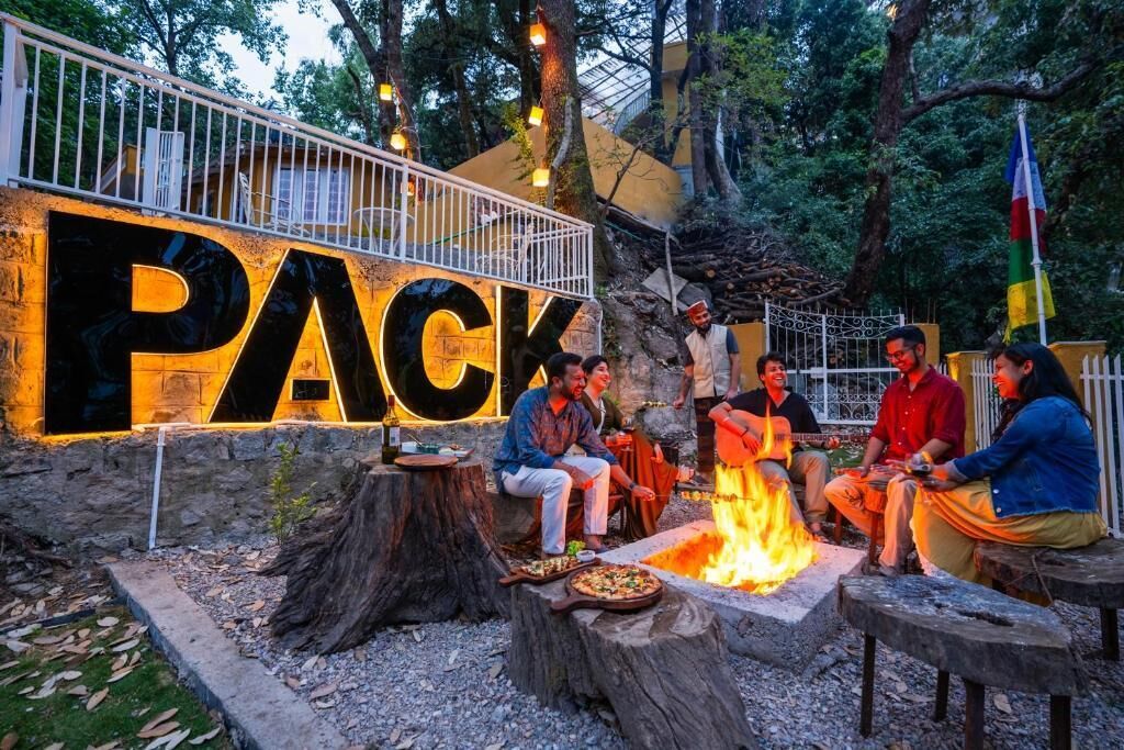 A group of people sitting around a campfire in an outdoor setting with a large illuminated sign that reads "PACK".