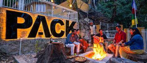 A group of people sitting around a campfire in an outdoor setting with a large illuminated sign that reads "PACK".