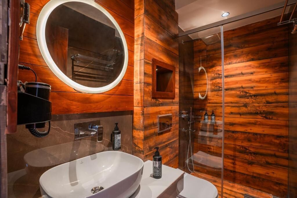A modern bathroom with a large oval mirror, wooden paneling, a white sink, and a glass-enclosed shower area.
