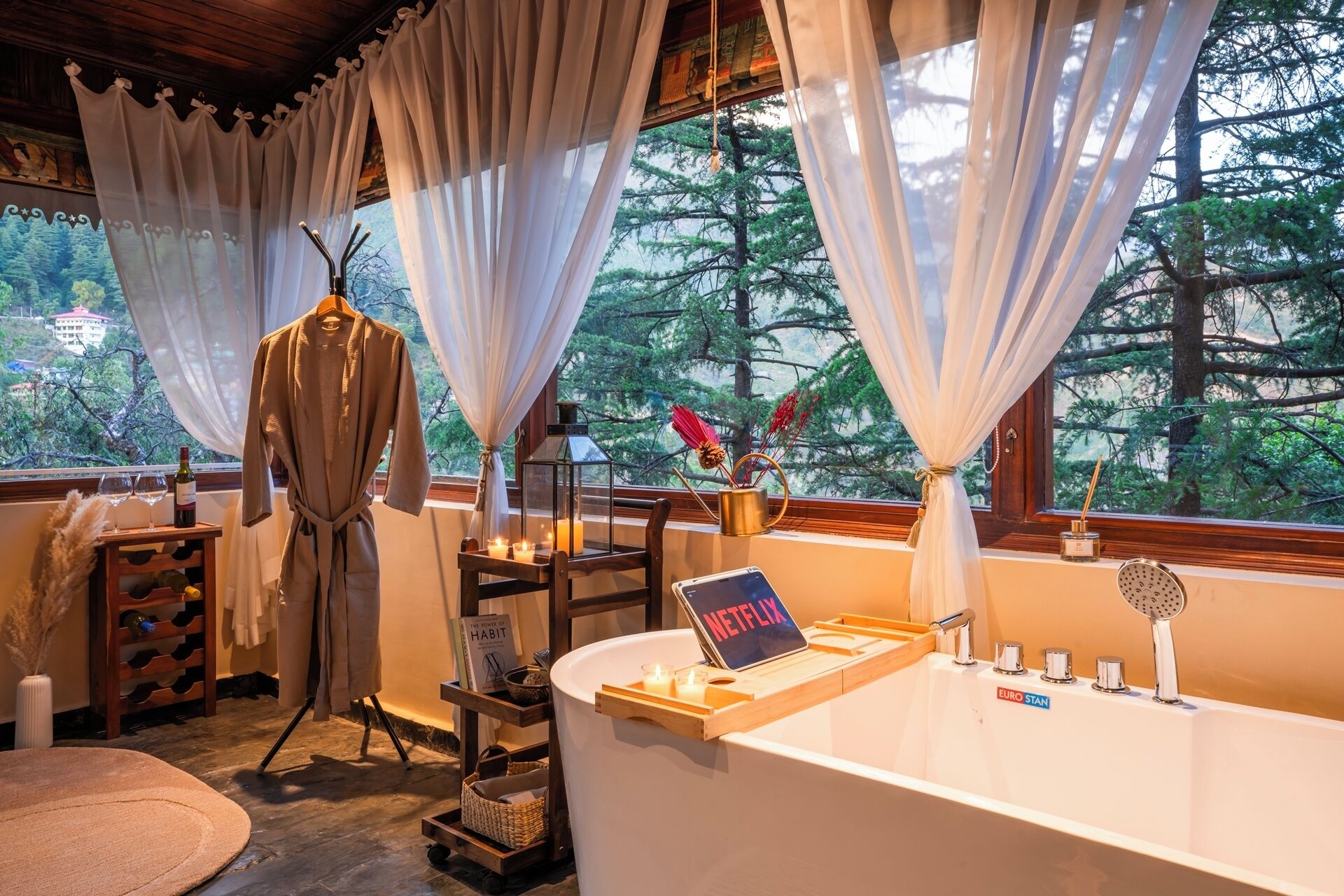 A bathroom with a bathtub, a vanity with a mirror, a sink, and a window with curtains. There is a robe hanging on a rack, a tray with candles, and a book titled "The Power of Habit" on a shelf.