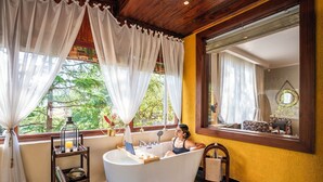 Ein Badezimmer mit einer Badewanne, einem hölzernen Regal mit Büchern, einem Fenster mit Vorhängen und Blick ins Grüne.