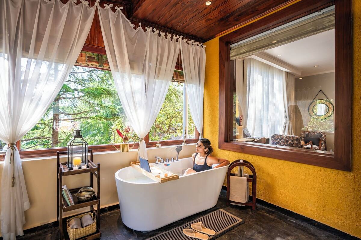 Ein Badezimmer mit einer Badewanne, einem hölzernen Regal mit Büchern, einem Fenster mit Vorhängen und Blick ins Grüne.