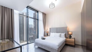 Ein modernes Hotelzimmer mit einem großen Bett, zwei Nachttischen mit Lampen, einem Glastisch und Blick auf hohe Gebäude durch große Fenster.
