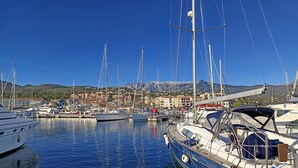 Un puerto deportivo con varios veleros amarrados, un pueblo al fondo y un paisaje montañoso.