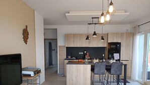 Una cocina moderna con armarios de madera, una isla central e iluminación colgante. Hay un comedor con sillas y un televisor en la sala de estar.
