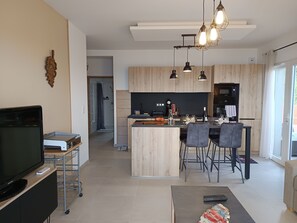 Eine moderne Küche mit Holzschnitzschränken, einer zentralen Kochinsel und Pendelleuchten. Im Wohnzimmer befindet sich ein Essbereich mit Stühlen und ein Fernseher.