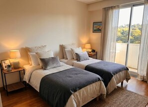 A bedroom with two beds, bedside lamps, a view of trees through a window, and a framed picture on the wall. - Panoramic Sea-View Apartment w terrace in Cannes, 2 Bed, 2 Bath Pool & Parking (Cannes)