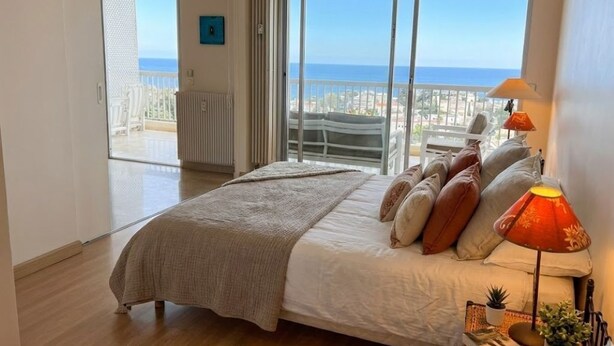 Une chambre avec un grand lit, une table de chevet, une lampe et une vue sur la mer, accessible par une porte-fenêtre donnant sur un balcon.