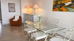 Un coin repas avec une table en verre et des chaises en métal blanc, un fauteuil marron et un tableau accroché au mur.