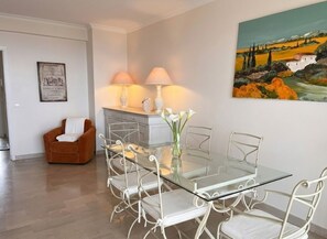 A dining area with a glass table and white metal chairs, a brown armchair, and a painting on the wall. - Panoramic Sea-View Apartment w terrace in Cannes, 2 Bed, 2 Bath Pool & Parking (Cannes)