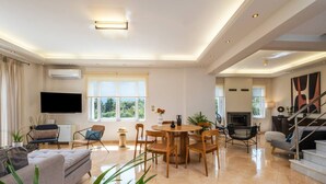 Uma sala de estar espaçosa com uma televisão grande, um espaço de refeições com mobiliário de madeira e uma escada que dá acesso a um andar superior.