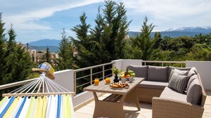 Uma varanda com mobiliário de vime, uma mesa com frutas e bebidas, e vista para montanhas e árvores.