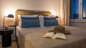 Uma cama bem arrumada com um livro, um chapéu de palha, duas almofadas estampadas e abajures em um quarto de hotel com vista para o mar.