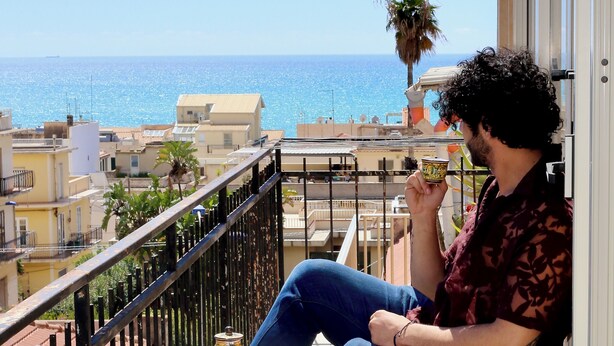 A balcony with a view of the sea, a person sitting, and a cup of coffee.