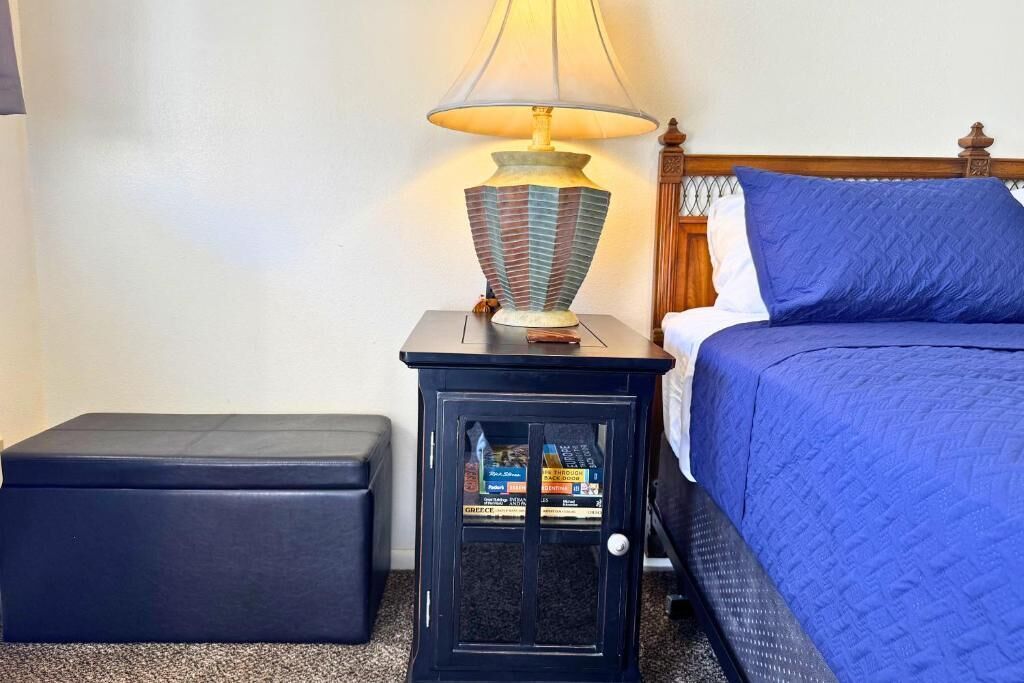 A neatly made bed with a blue quilt, a bedside lamp, and a small cabinet with books.