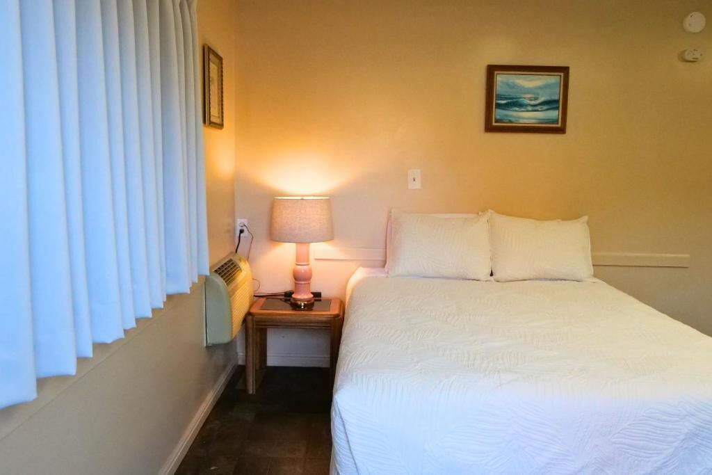 A hotel room with a bed, a nightstand with a lamp, a painting on the wall, and a window with blinds.