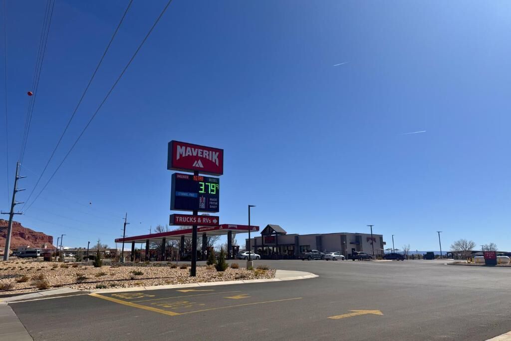 Una estación de servicio Maverik bajo un cielo azul despejado, un cartel con los precios de la gasolina y un área de estacionamiento con espacios delimitados.