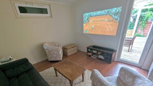 Ein Wohnzimmer mit einem großen Bildschirm, der ein Tennismatch zeigt, einer Couch, einem Couchtisch und einem gemusterten Sessel.