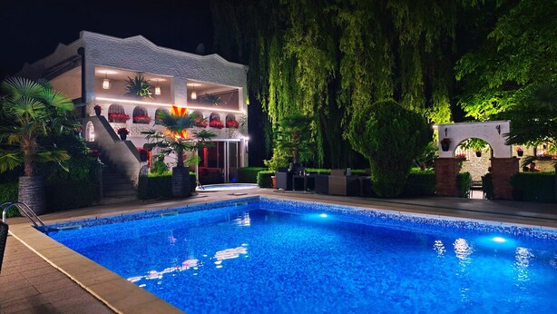 A swimming pool with a two-story building in the background, surrounded by greenery and a stone wall.
