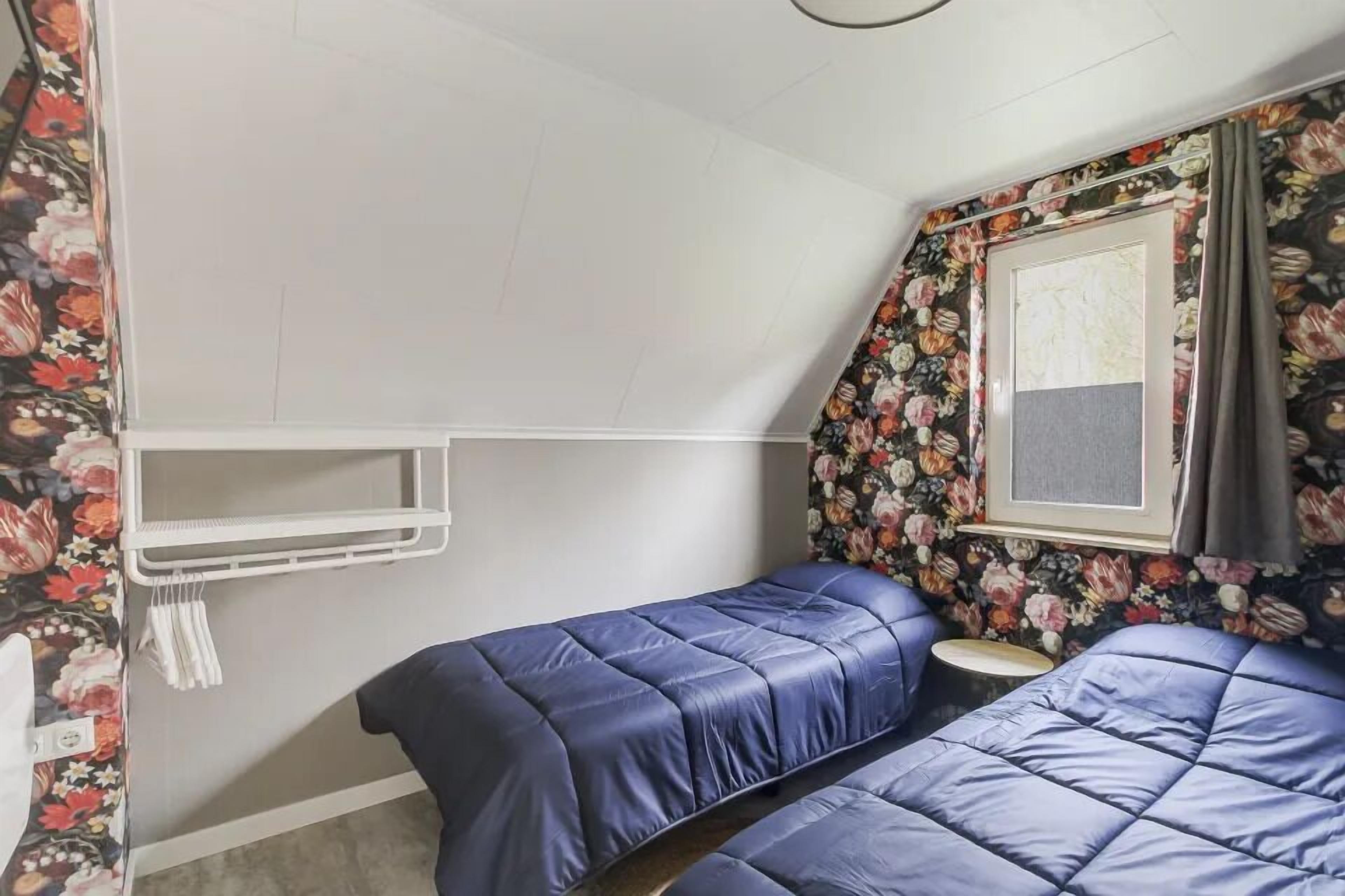 A small room with two beds, a window with a floral curtain, and a white shelf.