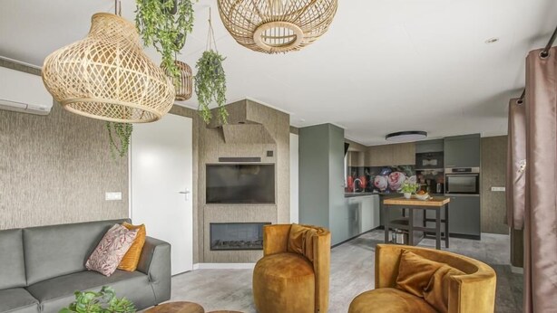 Un salon moderne avec un canapé gris, une table basse ronde en bois et deux fauteuils de couleur moutarde. Un téléviseur est fixé au mur, on aperçoit une cuisine au fond et une plante suspendue.