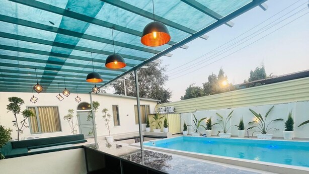 A modern outdoor kitchen with a built-in barbecue grill, a pool, and a covered patio area with hanging lights. - Olivine By Housiify (Gurugram)