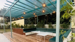 An outdoor patio with a wooden deck, a green cushioned seating area, and hanging pendant lights. - Olivine By Housiify (Gurugram)